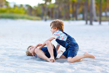 Two little kids boys having fun on tropical beach, happy best friends playing, friendship concept. Siblings brothers, twins fighting, running and jumping in family look with palms trees on background.