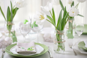 Decor and table setting of the Easter table with white tulips and dishes of green and white color. Easter decor in the form of Easter bunnies  green color with white polka dots.