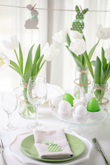 Decor and table setting of the Easter table with white tulips and dishes of green and white color. Easter decor in the form of Easter bunnies  green color with white polka dots.