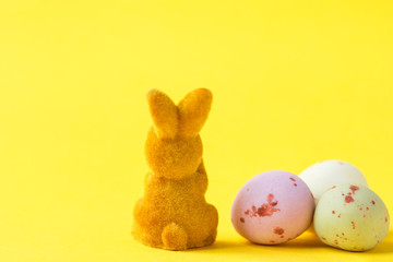 Fluffy brown toy Easter bunny sitting with his back to the viewer on yellow background with bunch of multicolored pastel chocolate eggs. Holiday banner poster greeting card template. Creative