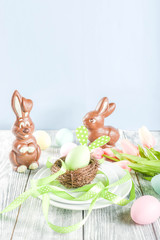 Easter holiday table setting with rabbits and eggs, plates with colorful eggs and chocolate bunny rabbits, festive ribbon, wooden background top view copy space