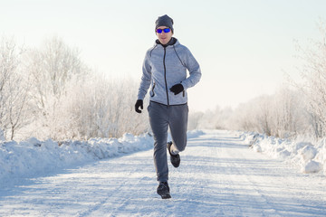 Young, adult alone man in tracksuit, hat and sunglasses running on snowy road outside of city. Enjoying sport in sunny winter morning. Daily active lifestyle. Life motion. Front view.