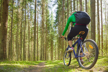 Young adult woman cycling through green forest in fresh air. Backpack on back. Enjoying sport in sunny day. Outdoor activities on weekends. Active recreation in nature. Low angle view.