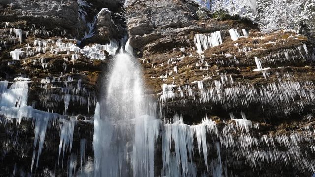 NATURE BACKGROUND, SLOW MOTION 4K Close up of a stunning waterfall falling from the side of a mountain cliff covered in icicles on sunny winter day. Waterfall falling past numerous white icicles froze