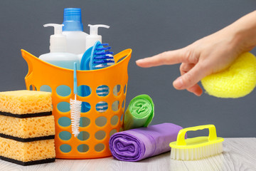 Bottles of dishwashing liquid, brush in a basket and sponges, garbage bags with female hand on gray background.