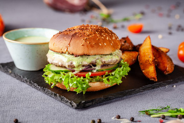 American burger with meat patty, tomato, cucumber, green sauce. Sauce and for french fries on a black stone slate board. Dish in a fast food restaurant. Copy space