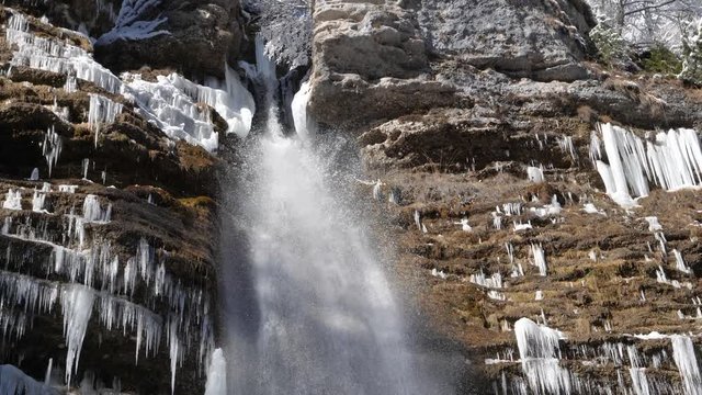 NATURE BACKGROUND, SLOW MOTION 4K Close up panning shot from top to bottom of a waterfall on a winter day surrounded by frozen icicles. Waterfall falling past numerous white icicles frozen in winter w