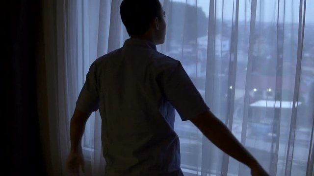 man opening the window curtain and looking away to the city at dawn