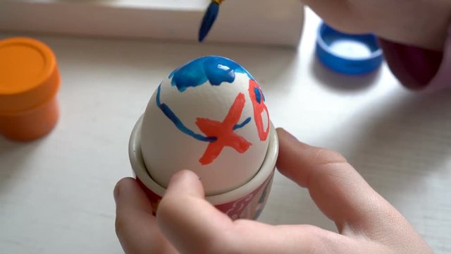 Extremely close-up, the child paints the Easter egg blue and red paint, sitting at the table. 4K. 25 fps