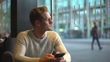 Slow motion attractive masculine european man sitting inside cafe gloomy, worried, looking outside window thoughtful space out, thinking, contemplating passersby, waiting reply phone message