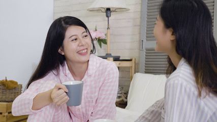 Two cheerful chinese women laughing while sitting comfortably in bed. young college girls roommates in pajamas lazy at home on weekend holidays. best female friends happy enjoy leisure time together