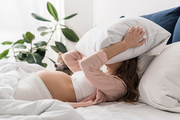 Pregnant woman lying in bed and holding pillow on head