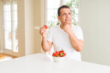 Middle age man eating strawberries at home serious face thinking about question, very confused idea