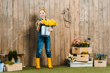 cheerful kid holding flowers in bucket near boxes with plants