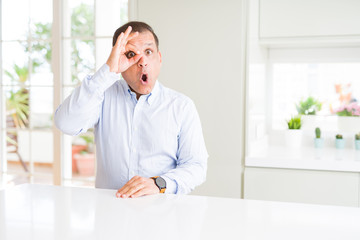 Middle age man sitting at home doing ok gesture shocked with surprised face, eye looking through fingers. Unbelieving expression.