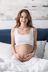 Cheerful pregnant woman sitting on bed and touching belly