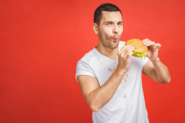 Funny hungry bearded man eating junk food. Excited young man greedily eating hamburgers isolated on red background.