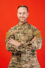 Young soldier in military wear keeping arms crossed and smiling  on red background.