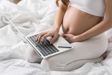 Naklejka premium Partial view of pregnant woman sitting on bed with laptop and credit card