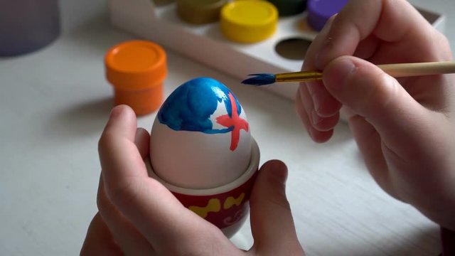 The child sits at the table and paints a brush Easter egg blue and red paint. Close up. 4K. 25 fps