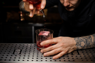 Bartender adding to the cocktail glass with ice cube a bitter from the dasher bottle