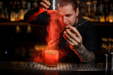 Professional tattooed bartender adding to an alcoholic cocktail a dried orange and powder