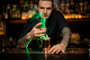 Bartender adding to a cocktail with dried orange a smoked cinnamon in the green light