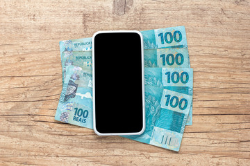 Banknotes of brazilian currency: Reais. Blank smartphone screen and cash bills on wooden table.