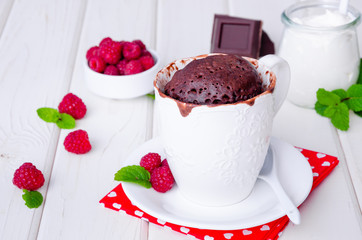 Chocolate mug cupcake with whipped cream, chocolate chips and fresh raspberries on a white wooden background.