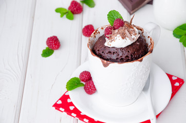 Chocolate mug cupcake with whipped cream, chocolate chips and fresh raspberries on a white wooden background.
