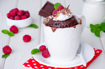 Chocolate mug cupcake with whipped cream, chocolate chips and fresh raspberries on a white wooden background.