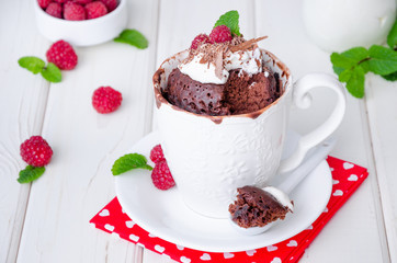 Chocolate mug cupcake with whipped cream, chocolate chips and fresh raspberries on a white wooden background.