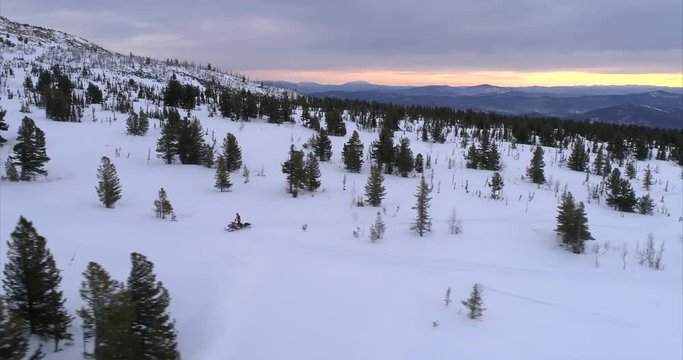 a guy on a mountain snowmobile rides on the background of a beautiful sunrise in a forest mountain valley with fields of snow. snowmobile traffic in the mountains. stock video footage. Aerial shot