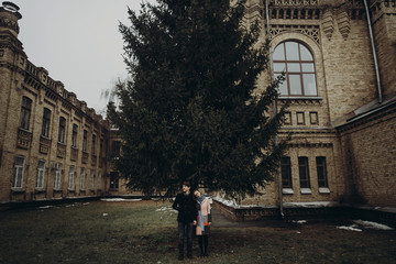Fototapeta premium stylish man and woman posing under tree. romantic calm atmospheric moment. couple hugging gently in autumn park at old building, showing true feelings. family togetherness concept