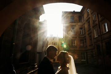 Beautiful blonde bride hugging handsome happy groom at sunset, newlywed couple kissing in old street, victorian architecture in the background