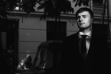 Handsome groom in black stylish suit with red tie posing outdoors at sunset, portrait of serious businessman in elegant clothing