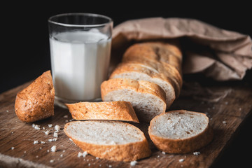 Healthy food. Long loaf of rural bread with two cut-off pieces lie on a wooden chopping board and a glass of fresh milk. Dark background.