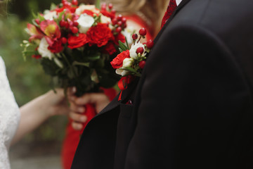 Handsome groom in stylish black suit holding wedding bouquet, white and red roses in fresh bouquet in man's hands outdoors