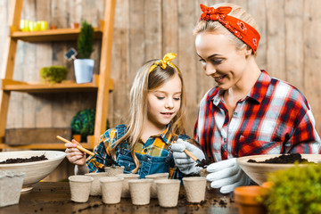 cheerful mother holding small shovel and putting ground in empty paper cups near cute daughter
