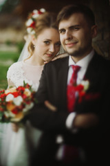 Happy newlyweds portrait, romantic couple, blonde bride with bouquet and handsome groom in stylish black suit posing outdoors, beautiful woman leaning against man