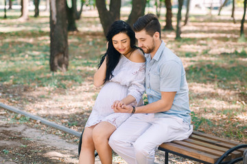 Fototapeta premium A young guy and his pregnant girl are sitting on a bench, against the background of greenery and trees. In anticipation of a child. First pregnancy I love children
