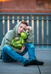 a young boy sits on a terrace on a wooden floor and hugs a large green frog in a golden crown
