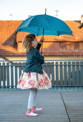 little girl in a floral dress and a blue jacket dances on the terrace with a blue umbrella