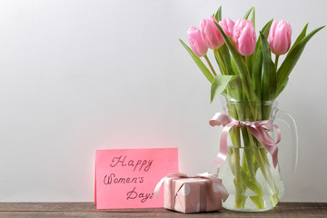 Beautiful bouquet of flowers of pink tulips in a vase and a gift box and the text of a happy women's day against a gray wall. place for text. Spring. holidays.