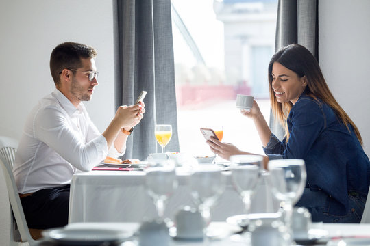 Young Hard-working Couple Using Their Mobile Phone In The Dining Room Of The Hotel.