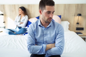 Attractive young couple ignoring each other after an argument sitting on bed at hotel room.