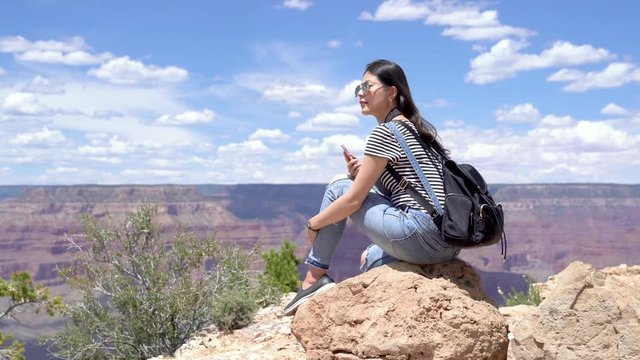 female explorer reading message on smartphone satisfied with mobile connection during hiking tour. girl backpacker sending beautiful desert view picture online sitting on viewpoint in Grand Canyon