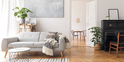 Modern scandianvian decor of living room with design sofa, elegant blanket, coffee table, plants and bookstand on the white wall. Design piano in the corner. Brown wooden parquet. View on dining room.