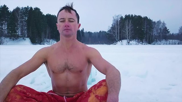 Young man in the red colorful pants meditates and tries to relax sitting on the ice of the lake with forest on the background. Fast dolly out camera (drone)