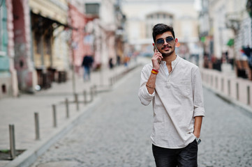 Stylish indian model man in casual clothes and sunglasses posed outdoor at street of India and speaking on mobile phone.